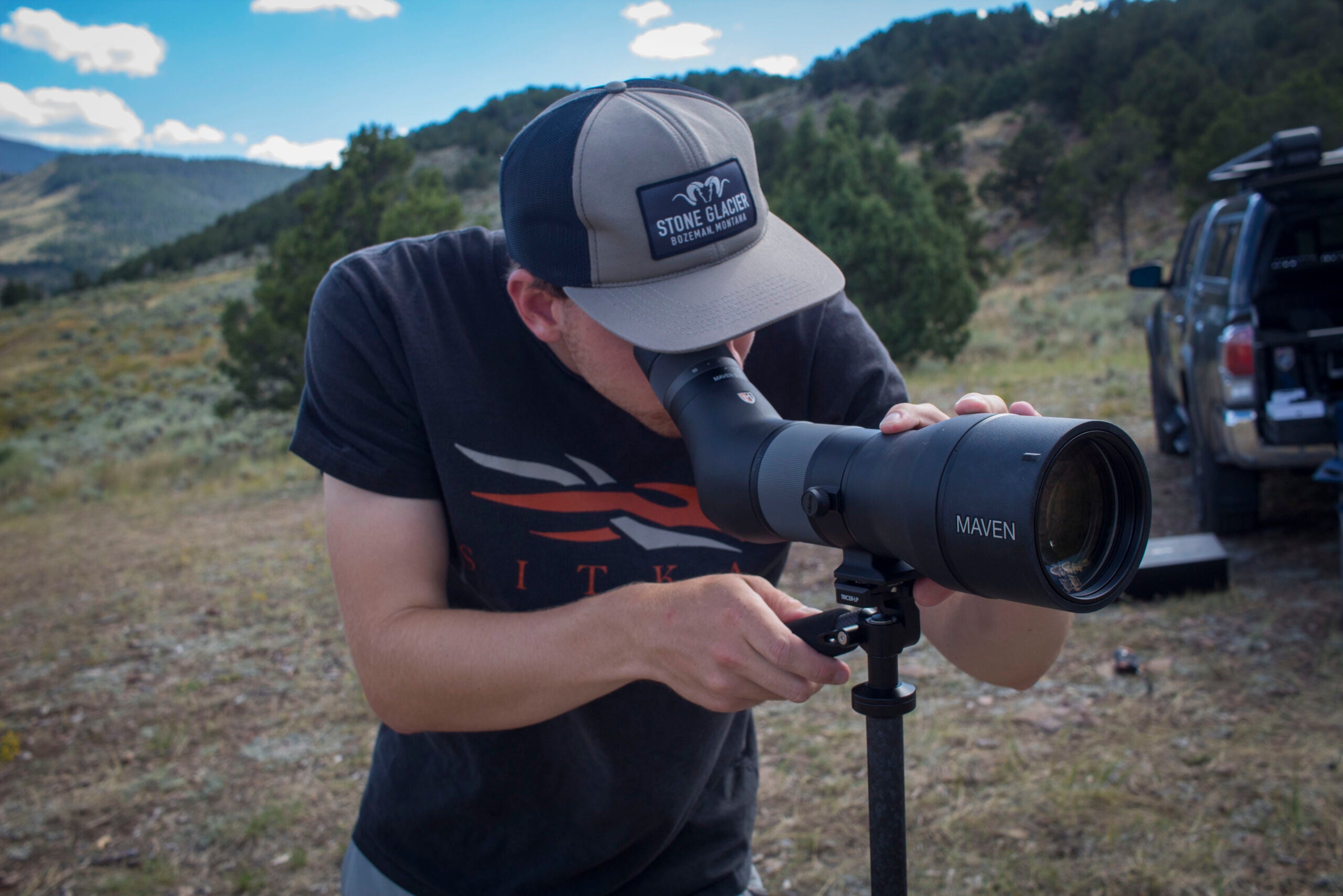 man looks through spotting scope with truck in the background