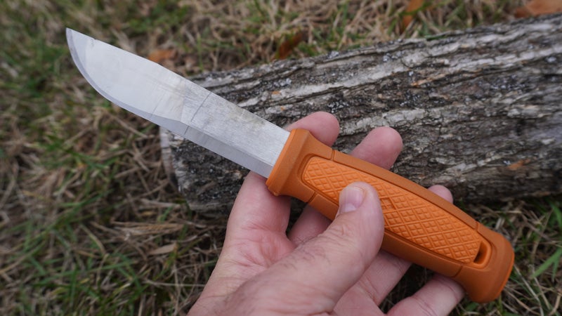 The tan and silver Morakniv Kansbol knife above a grey log and grassy lawn. 
