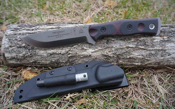 The red and black Tops Fieldcrafter fixed blade knife on a gray log on a lawn.