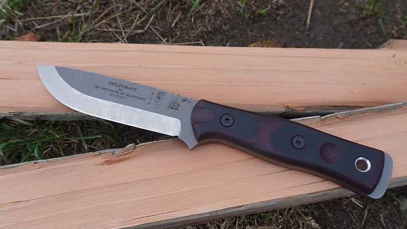 A silver, red, and black Tops Fieldcrafter fixed blade knife sitting on a split log on a grassy lawn. 