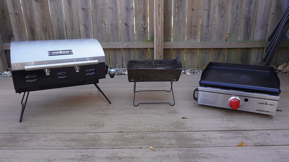 Three portable grills including a folding and griddle model sitting on a tan deck in front of a fence.