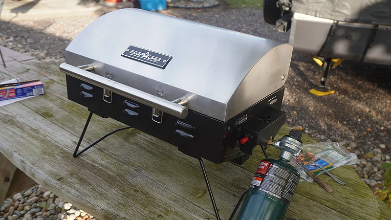 The Camp Chef portable bbq grill with a green propane tank inserted on a brown picnic table. 