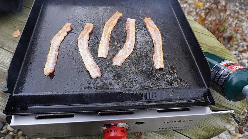 The black griddle of a Camp Chef Versatop stove with five slices of bacon simmering on the top. 