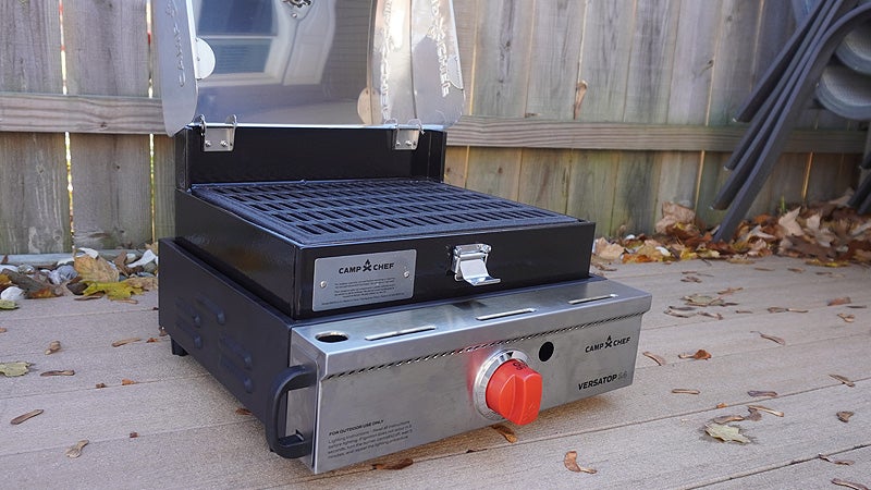 The black and silver Camp Chef 14-inch grill box sitting on top of the silver Camp Chef Versatop on a tan deck in front of a fence. 