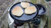 Three pancakes cooking on a griddle of a Coleman portable grill. 
