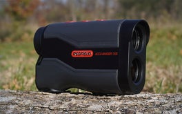 A gray, black, and red Redfield rangefinder sitting on a log with the lens facing right against a grassy background.