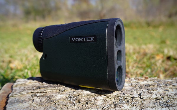 A Vortex Crossfire HD 1400 rangefinder sitting on a gray log in a grassy lawn.