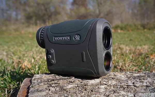 A green and gray Vortex Razor HD 4000 rangefinder sitting on a grey log on a grassy lawn.