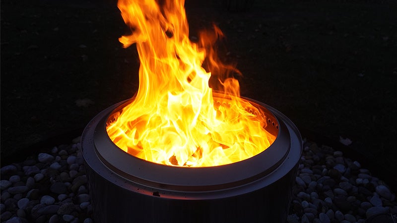 A large batch of flames licking out of the top of a Solo Stove Bonfire fire pit with rocks visible in the flickering light. 