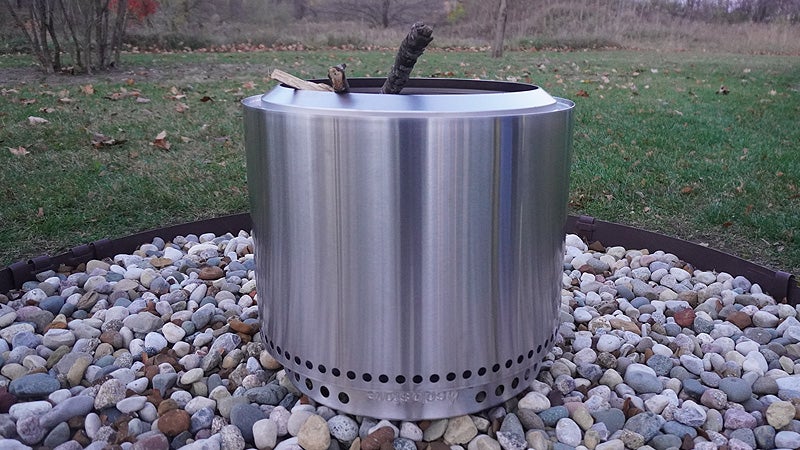 A silver Solo Stove bonfire with wood sticking out of the top while sitting on rocks in a grassy backyard. 