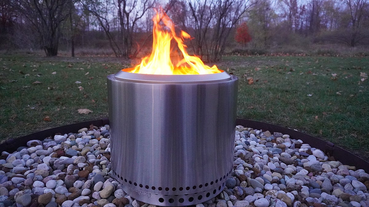 A silver Solo Stove Bonfire sitting on rocks in a backyard with orange flames licking from the top.