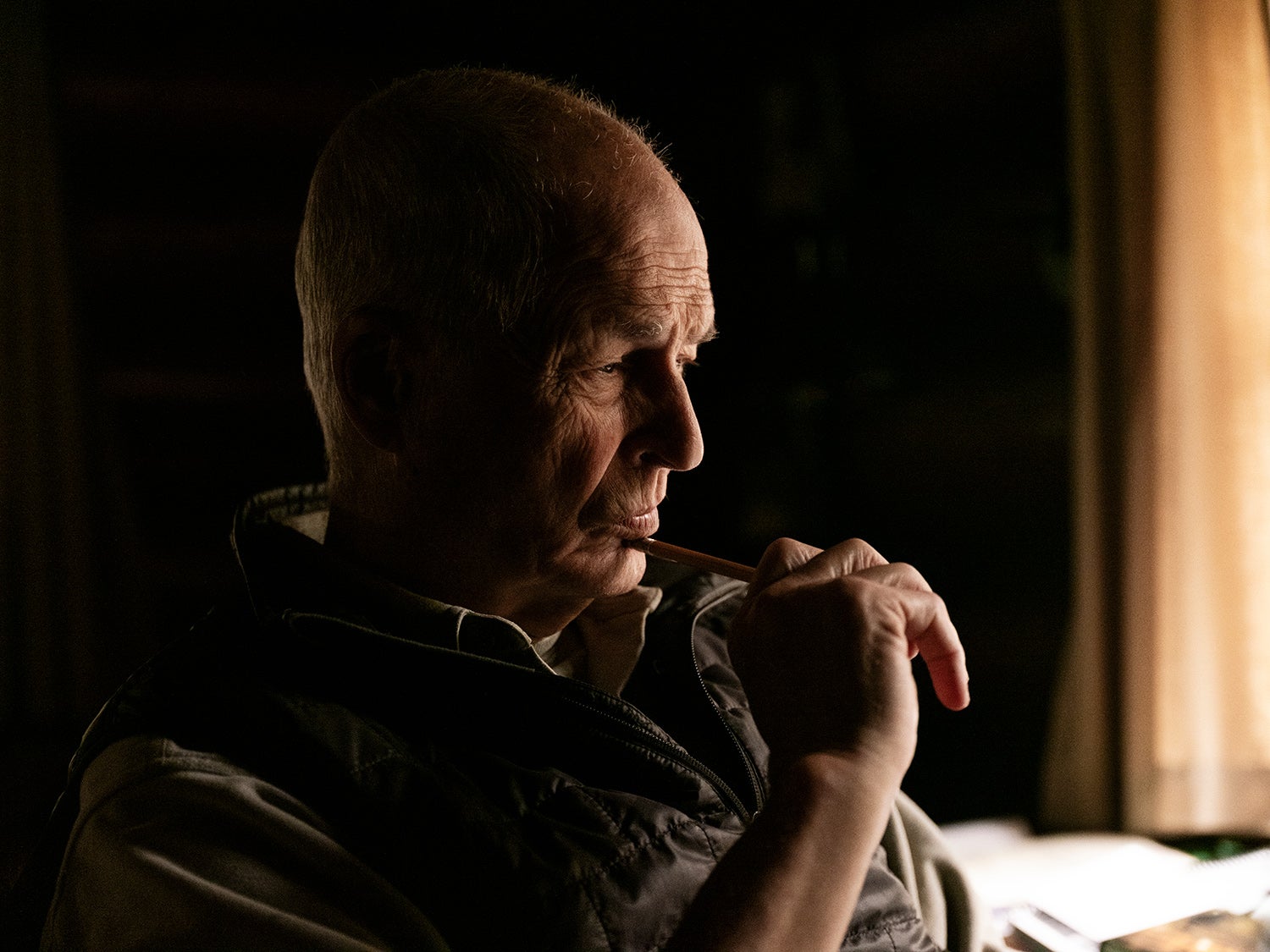 A man sits in a dark cabin, holding a pen and looking down at a desk.