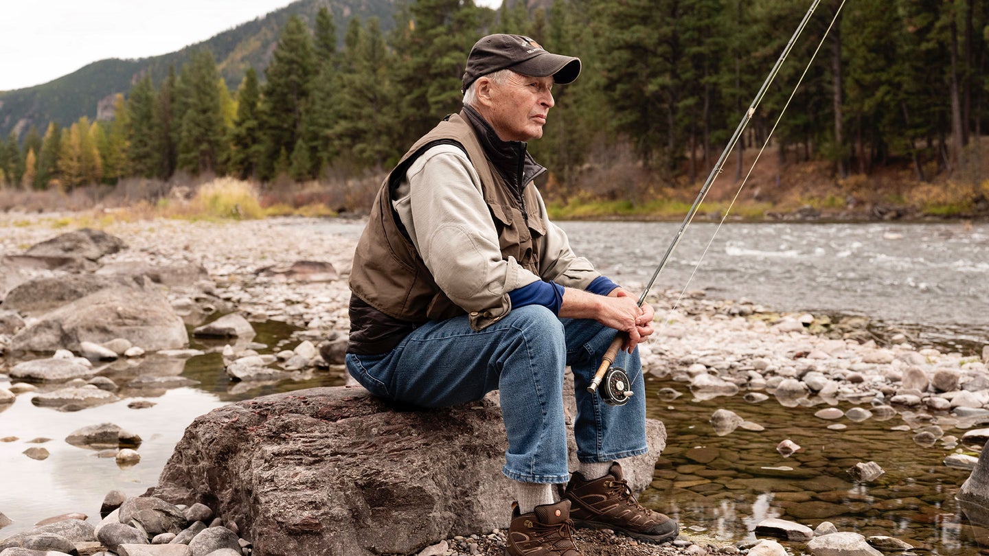 A fisherman dressed in jeans, a brown hat and a brown vest sits on a rock along a river with a fly fishing rod in his hand.