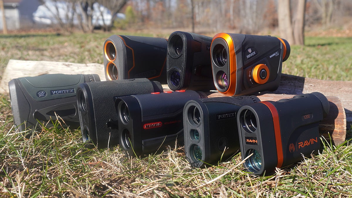 Eight rangefinders of varying brands and colors sitting on a log in a grassy lawn.