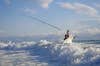 A fisherman stands in front of breaking wave with a fishing rod in the air.