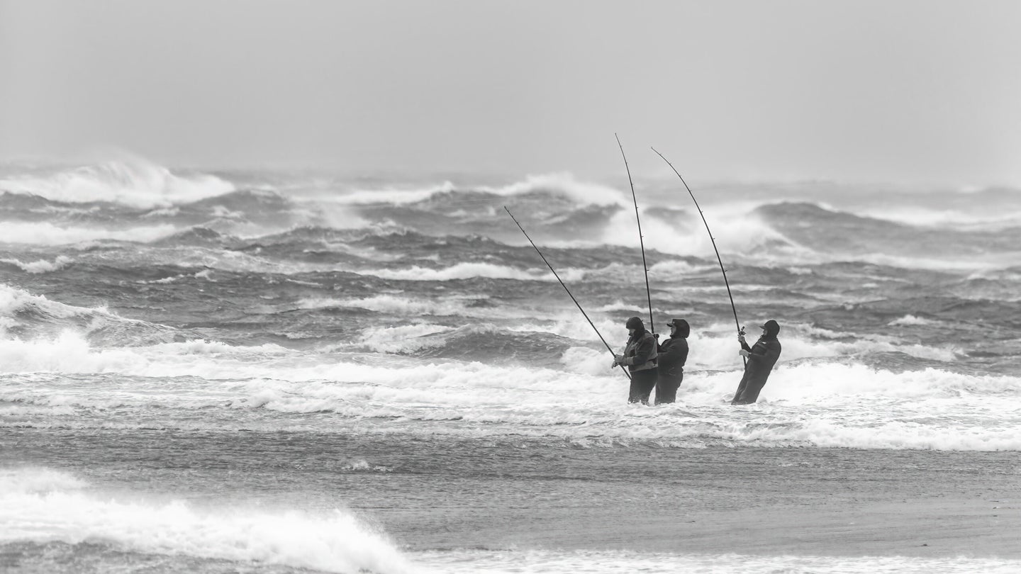 Three fishermen stand in the ocean with waves crashing while surf fishing