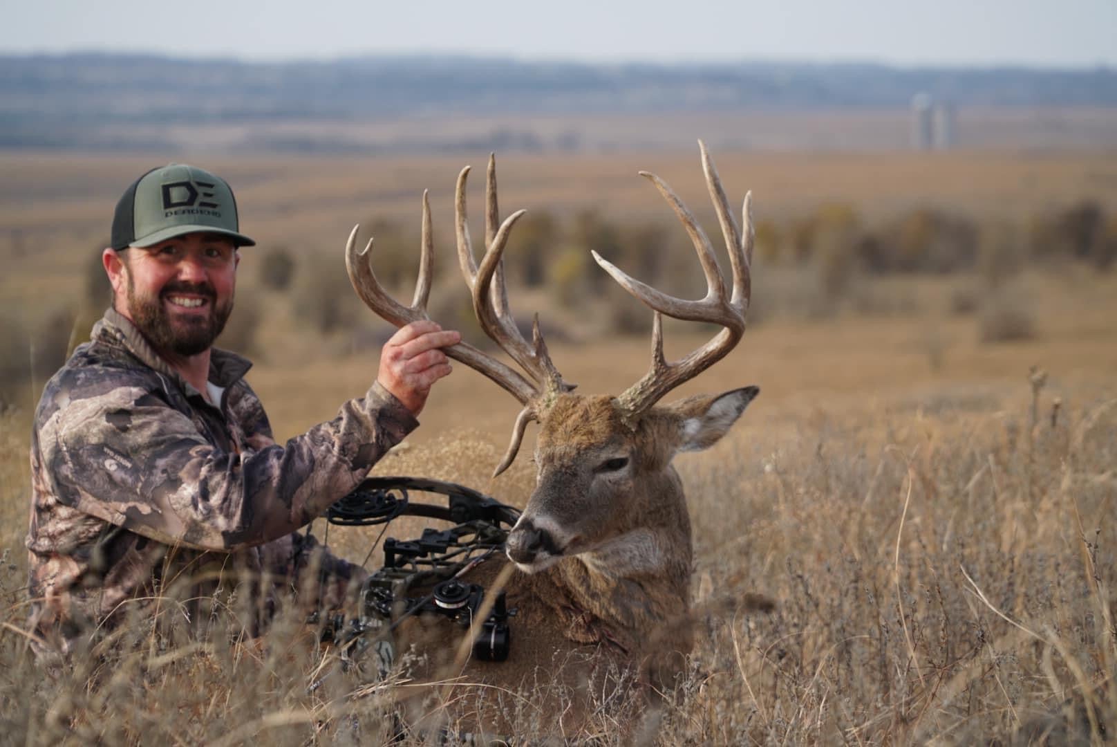 Hunter Patrick Paysinger sits in a grassy field, posing with a huge whitetail buck