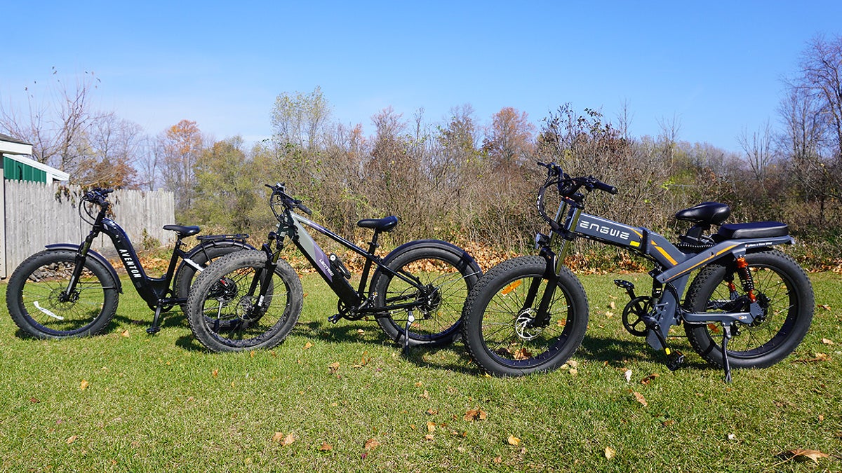 Three black and grey off-road electric bikes sitting in a grassy backyard with a blue sky overhead.