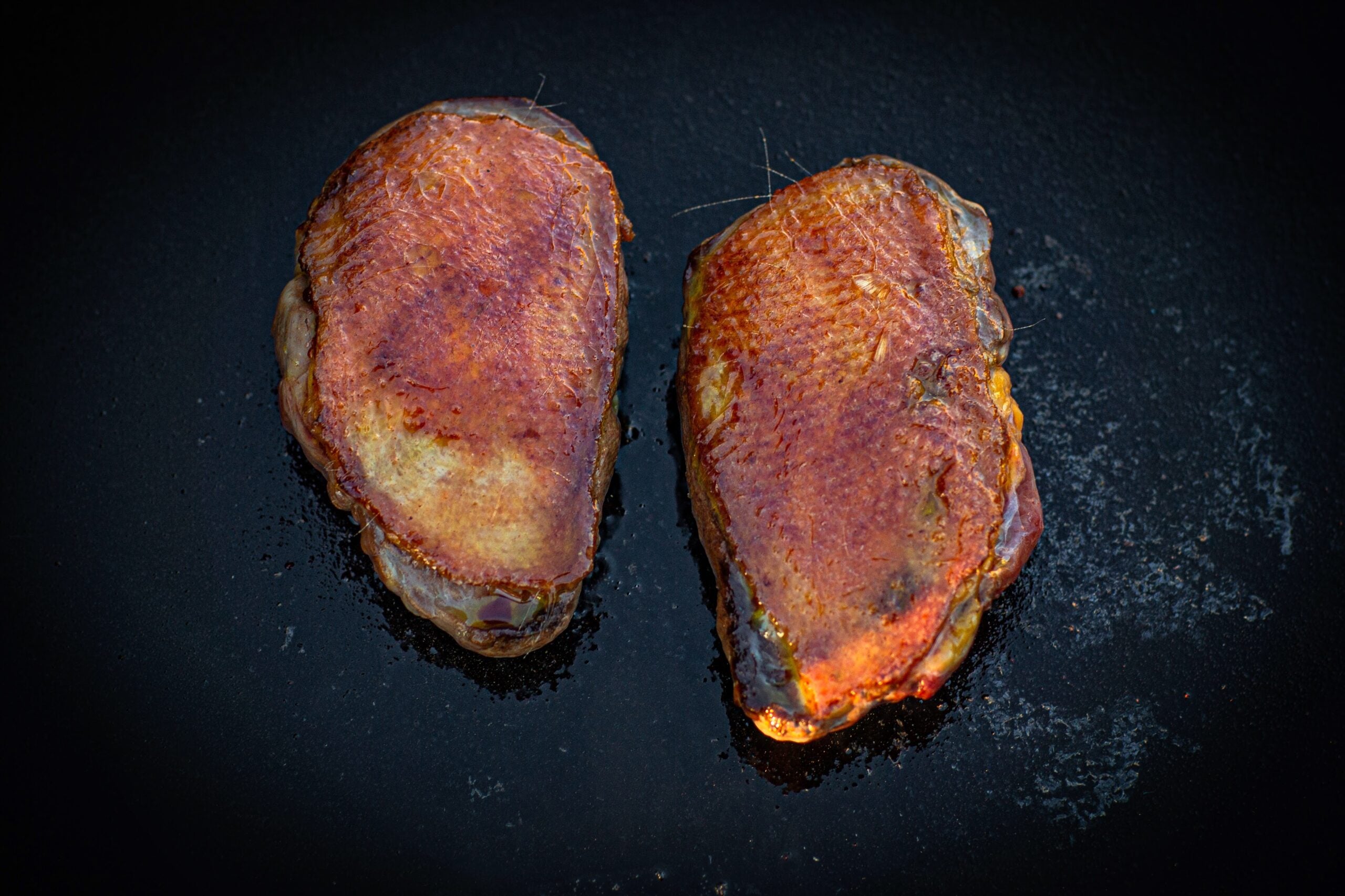 Two wild duck breasts cooking skin side up on a cast-iron surface