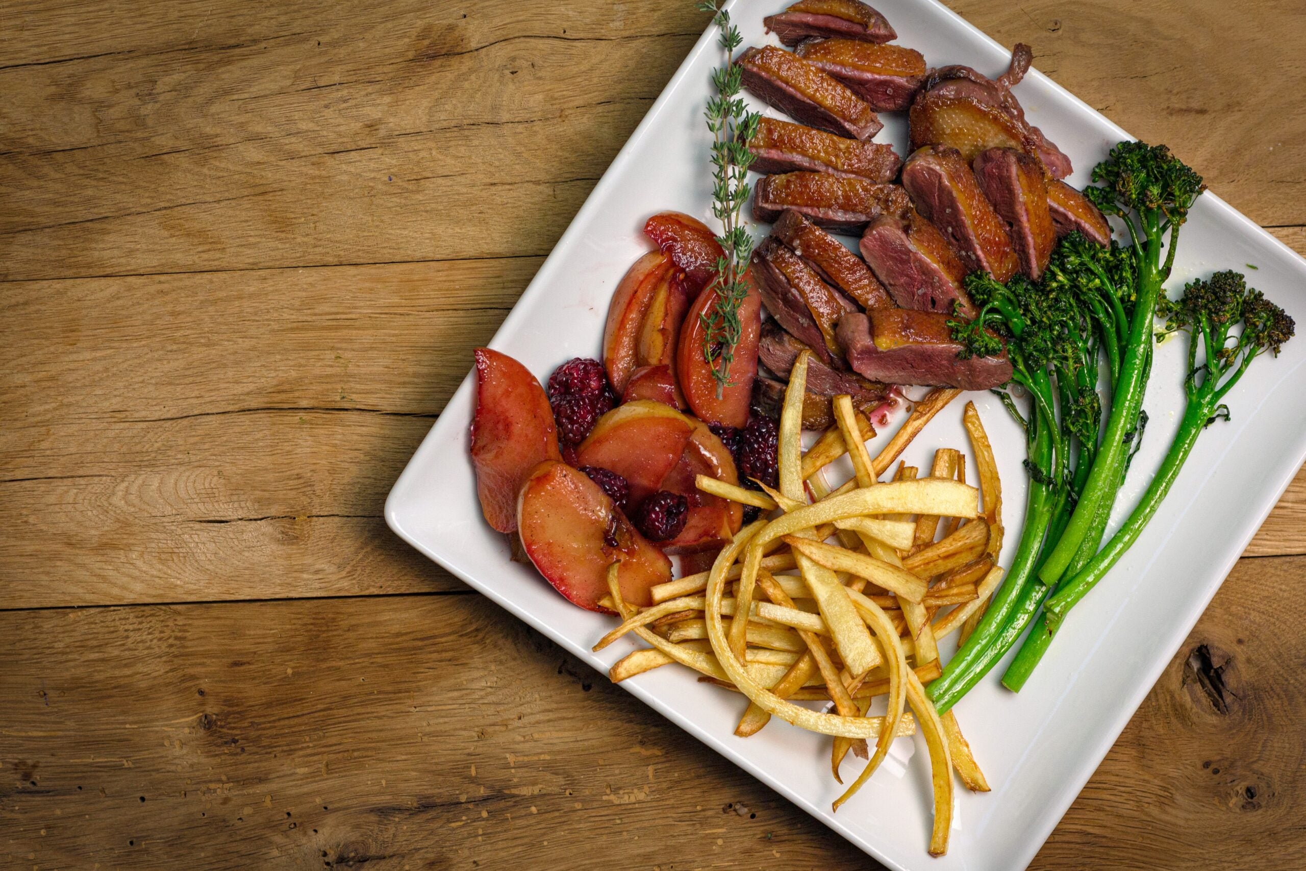 Sliced duck breast served alongside fries, broccoli rabe, and fruit on a white plate.