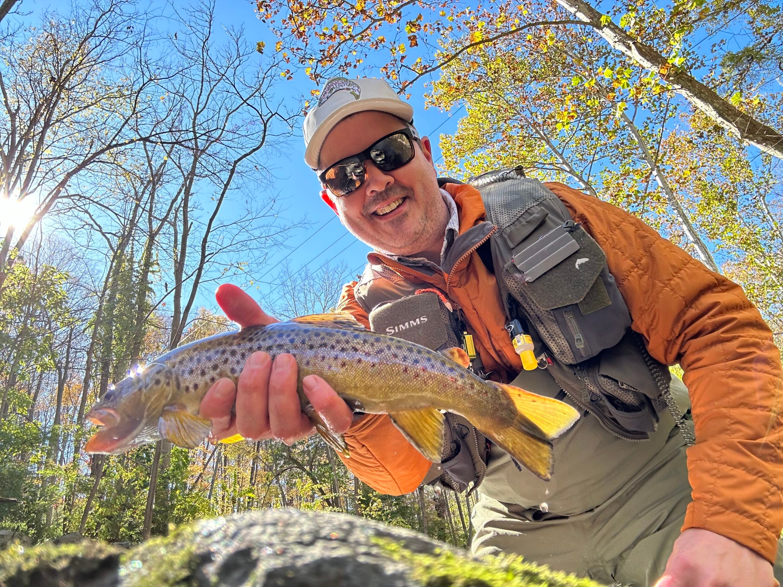 A fly fisherman in an orange jacket and fishing vest holds a brown trout