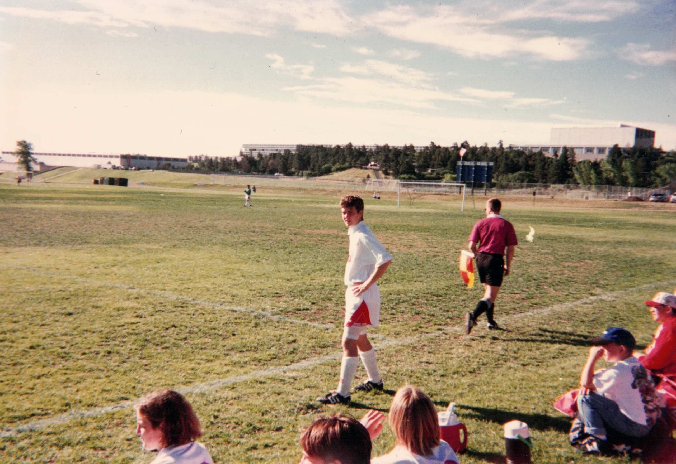 A young soccer player in a white uniform stands on the sidelines of a field.