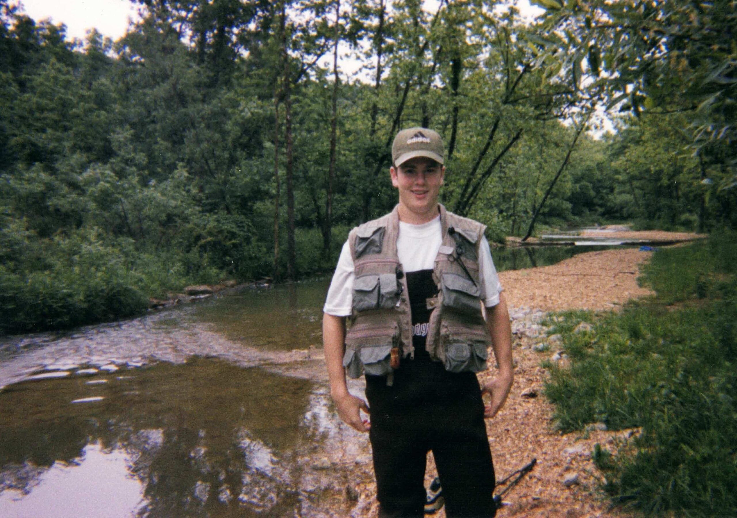 A young fisherman dressed in waders and a fishing vest stands on the bank of a river.