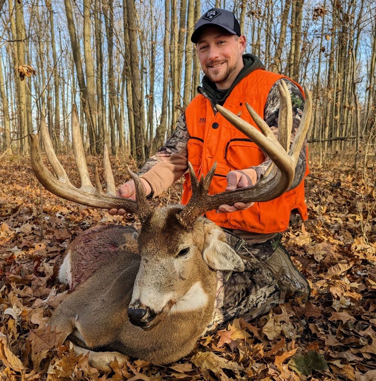Hunter in orange vest kneels on the ground in the woods, showing off a giant whitetail buck