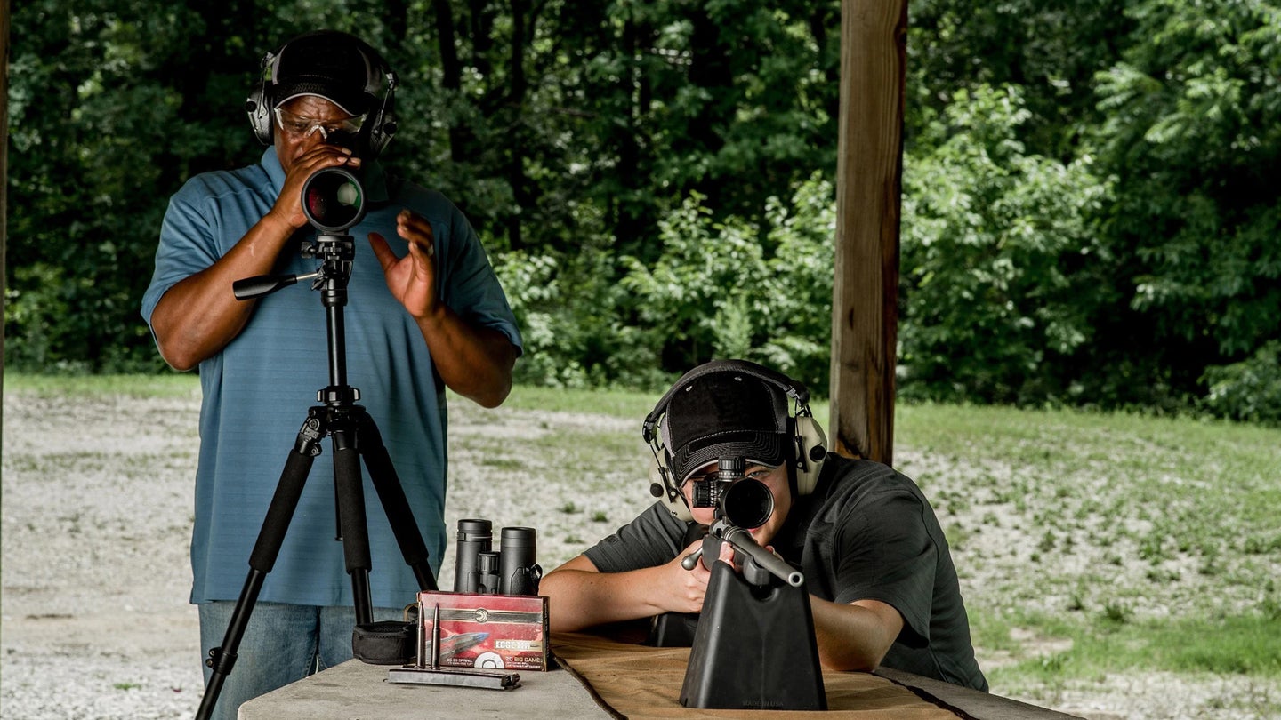 One man seated firing a rifle at a shooting range; another man looking downrange through a spotting scope