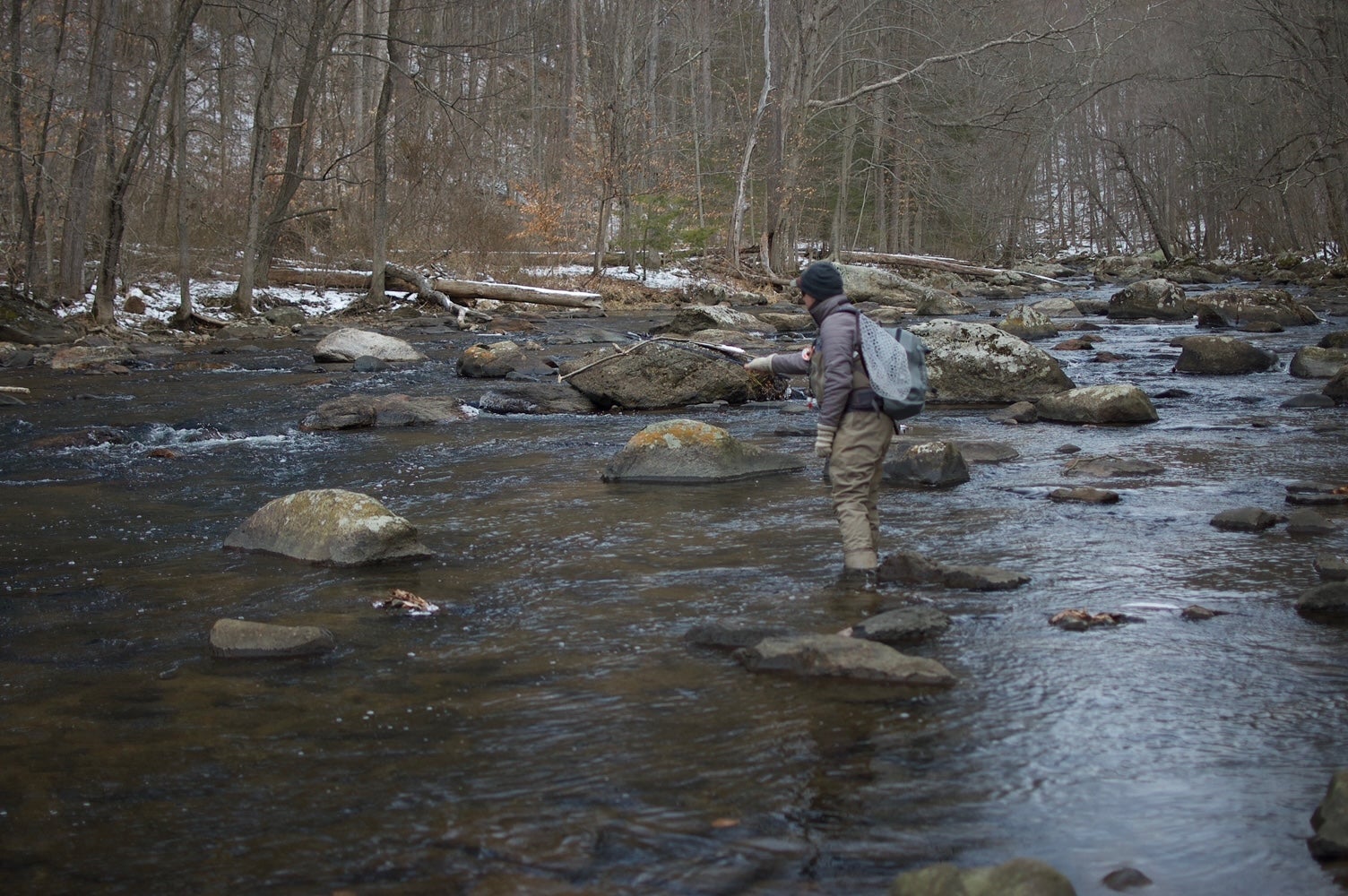 fly fisherman stands on boulder and makes cast into a riffle during the winter