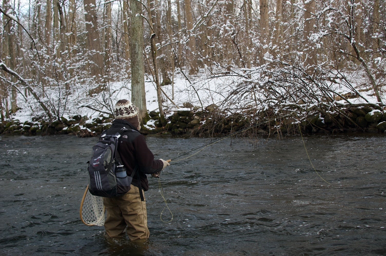 Fly fisherman stand in a river while making a cast with snow covered banks in the backgrond