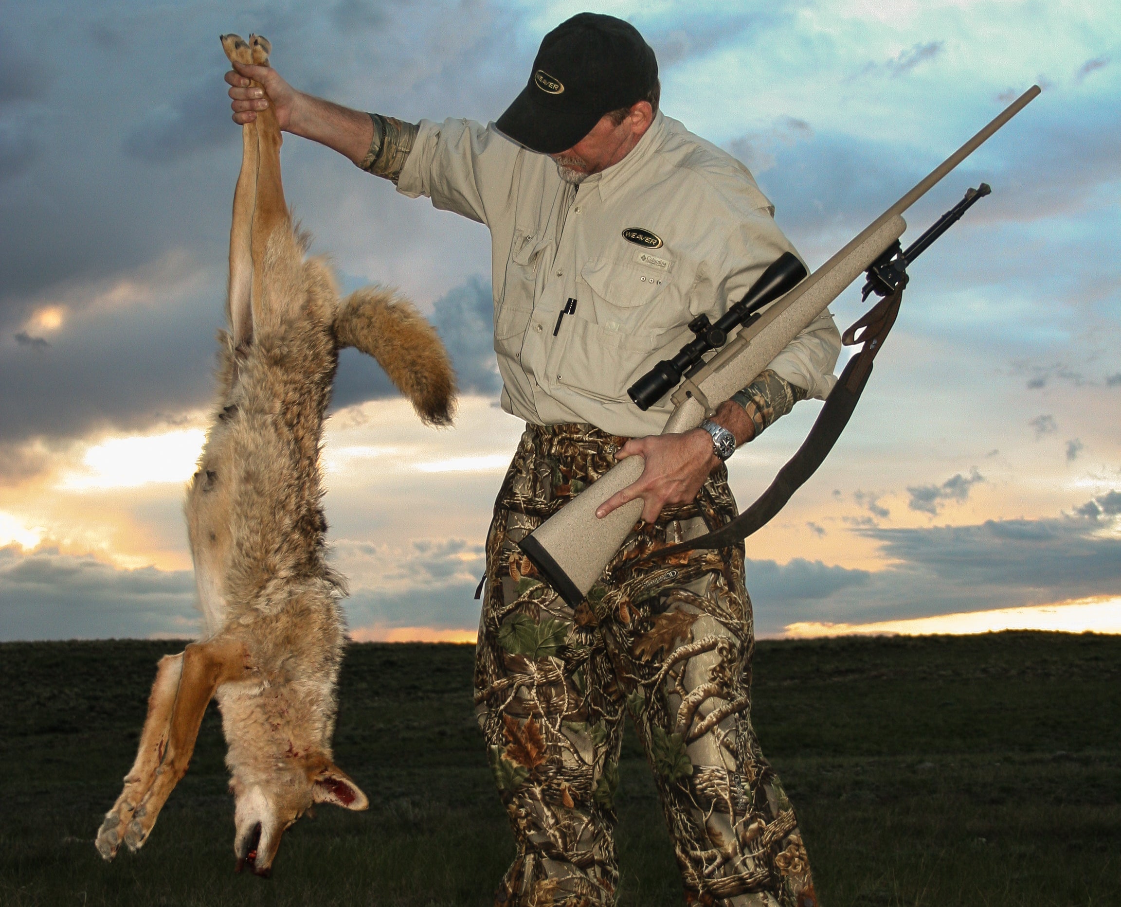 A hunter with a rifle in one hand hold up a coyote in the other