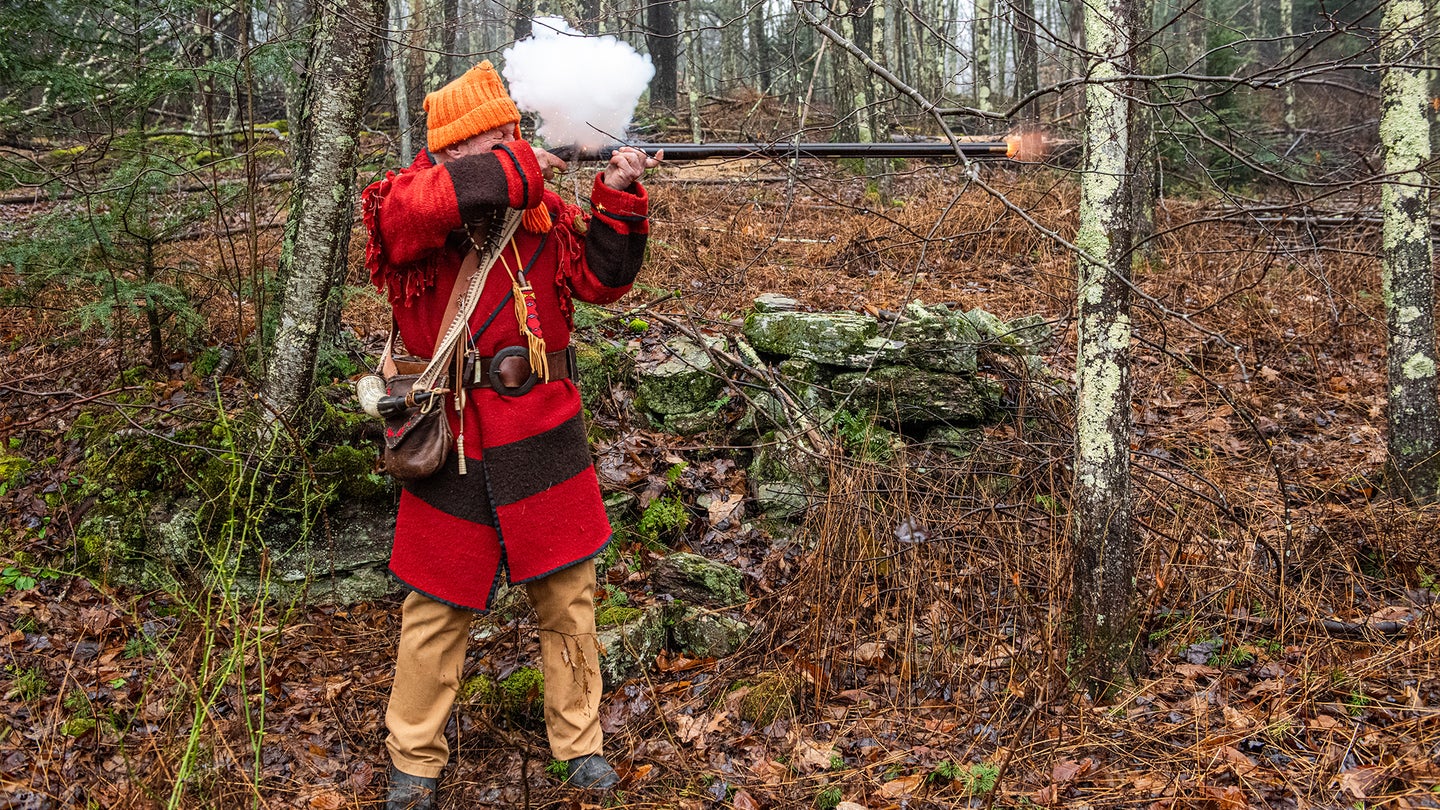Man wearing tradition muzzleloading garb fires a flintlock rifle, smoke billowing from the lock