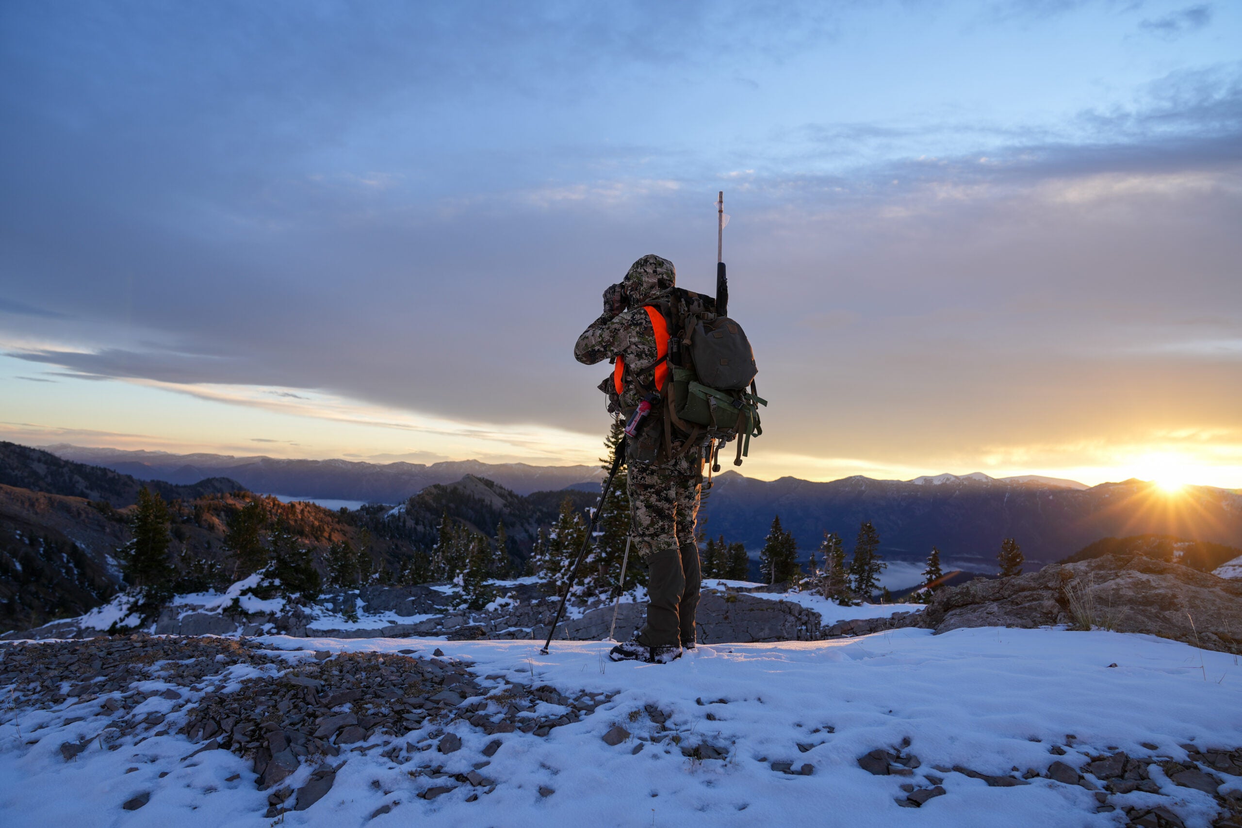 Hunter with rifle dressed in camo stands on the edge of a snowy mountain ridge looking for deer at sunrise in Wyoming