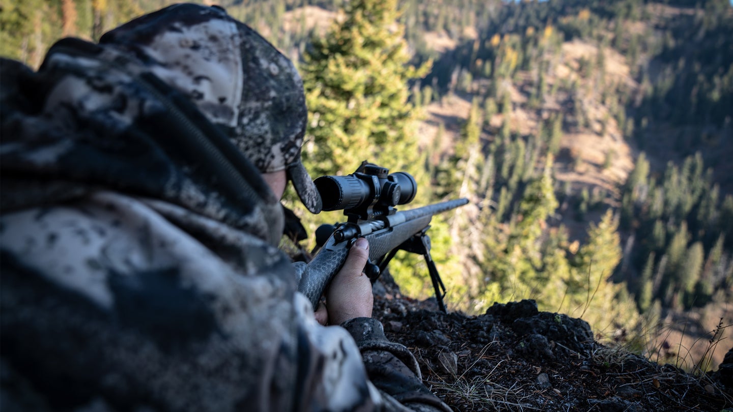 A long range hunter shoots a rifle from the prone position in a mountain setting