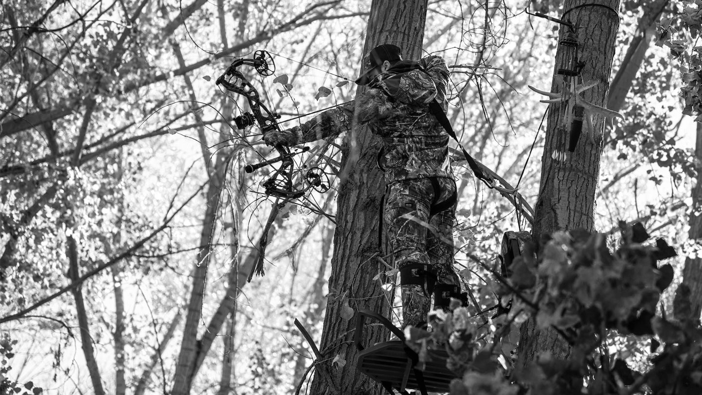 A bowhunter in a tree stand is at full draw with a bright sky behind him