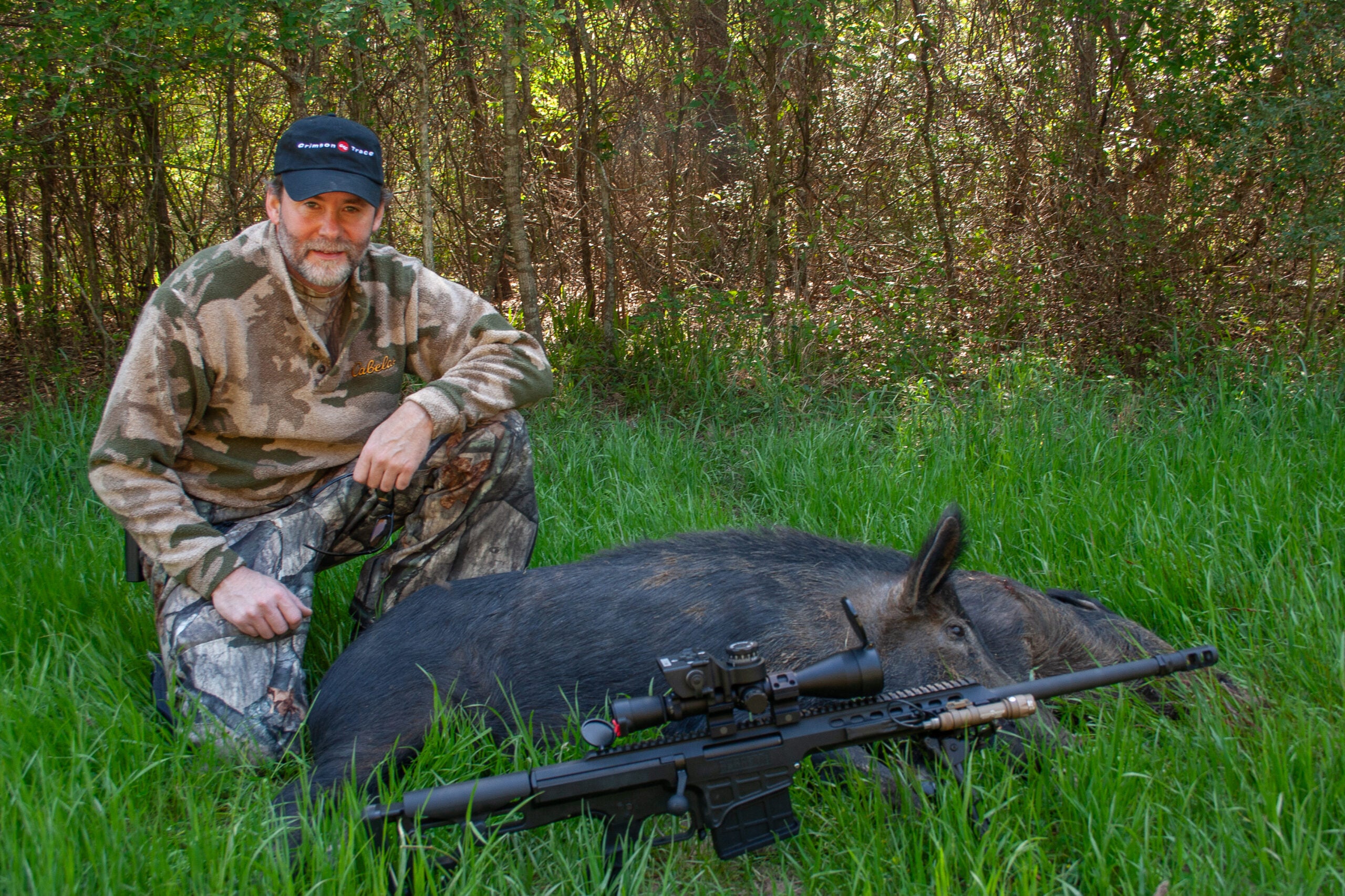 Hunter kneeling in the grass next to two wild hogs, with a 338 Lapua rifle in the foreground