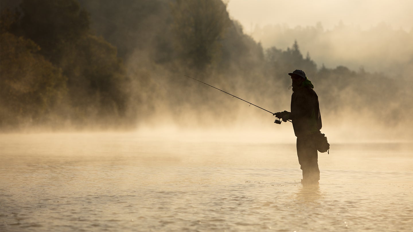 A man fishes while wading knee-deep in a river, surround by morning fog.
