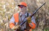 A hunter wearing blaze orange hat and vest holds a shotgun at port arms 