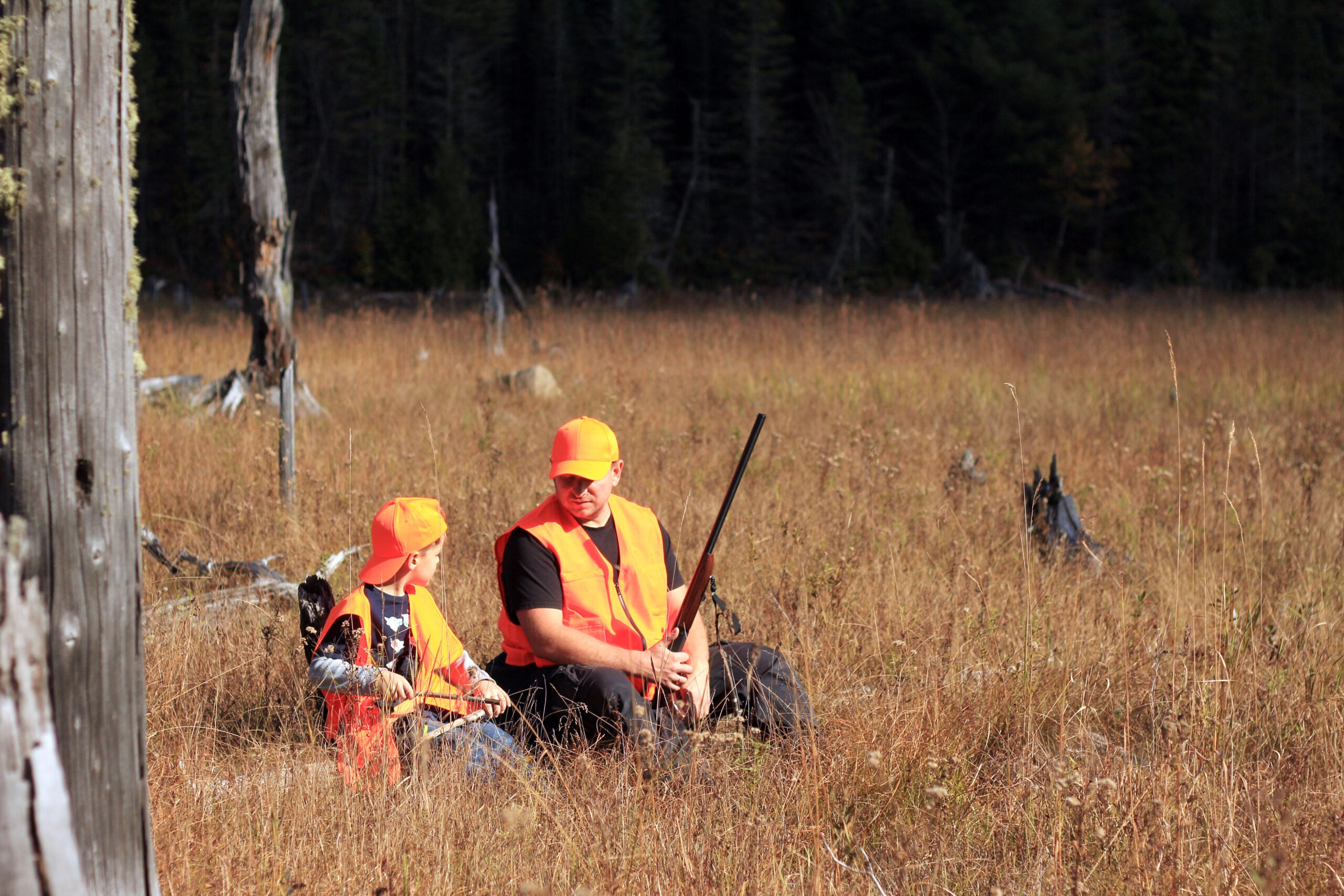 Father and son wearing blaze orange vests and hats sit on a log while hunting