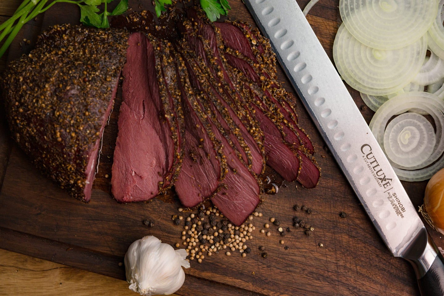 Slices of venison pastrami sit on a wooden cutting board next to a knife and garlic.