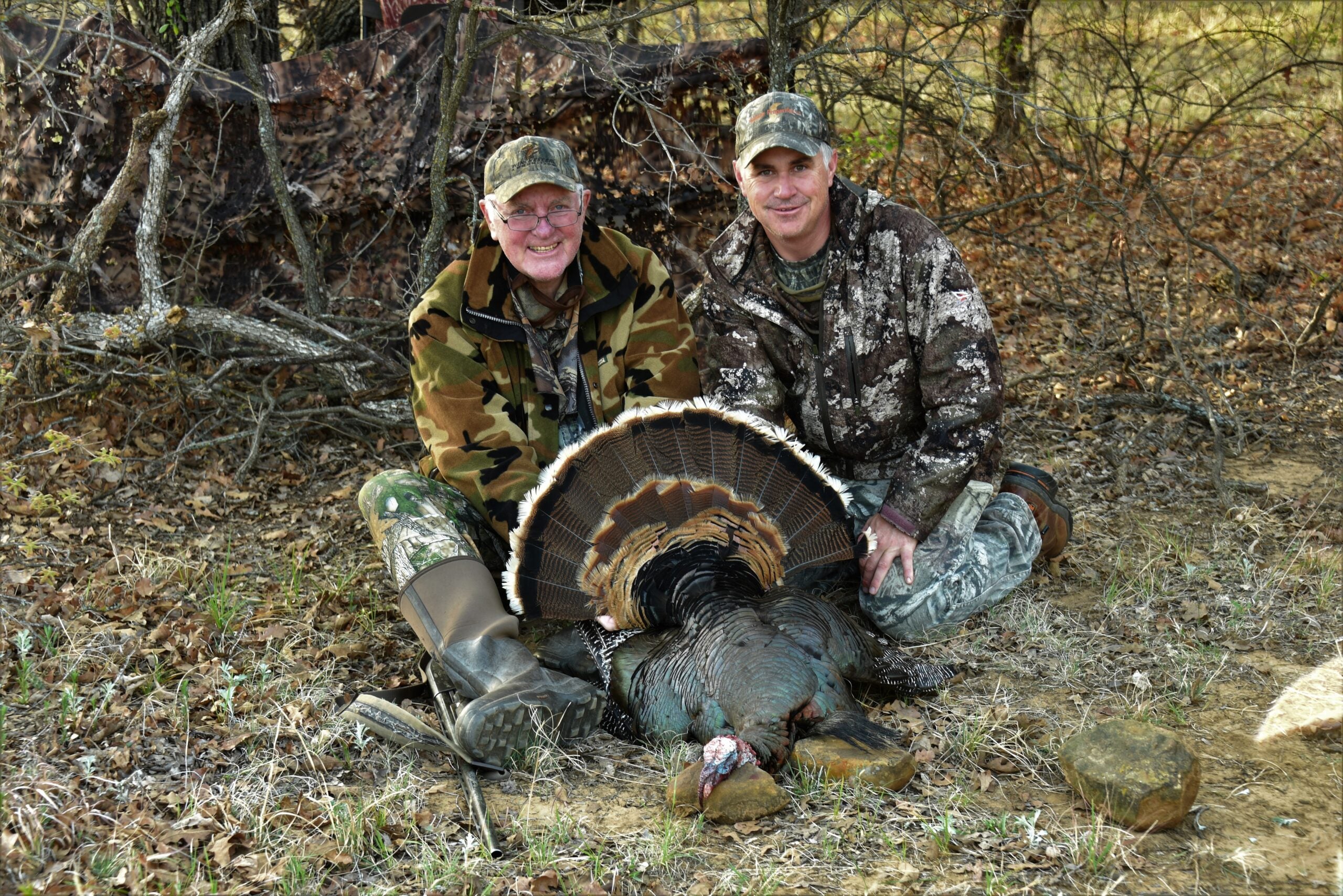 Two men dressed in camo sit on the ground showing off a harvested Rio Grande turkey