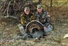 Two men dressed in camo sit on the ground showing off a harvested Rio Grande turkey