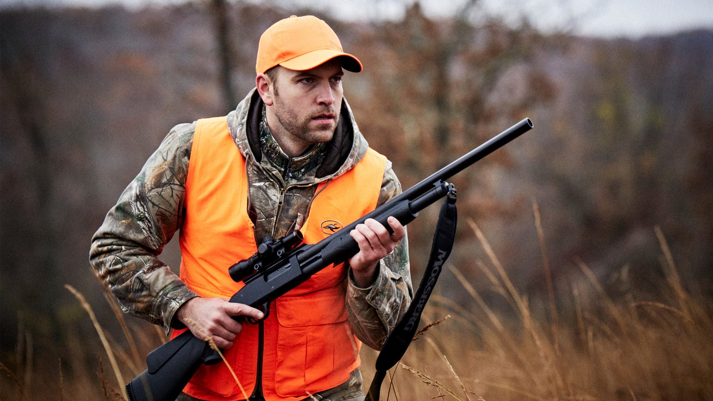Hunter in orange vest carrying a scoped shotgun and walking through grassy terrain