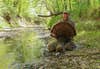 A hunter kneels along the edge of a wooded creek and shows off a tom turkey.