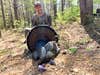 A hunter kneels on ground in the woods showing off a tom turkey.