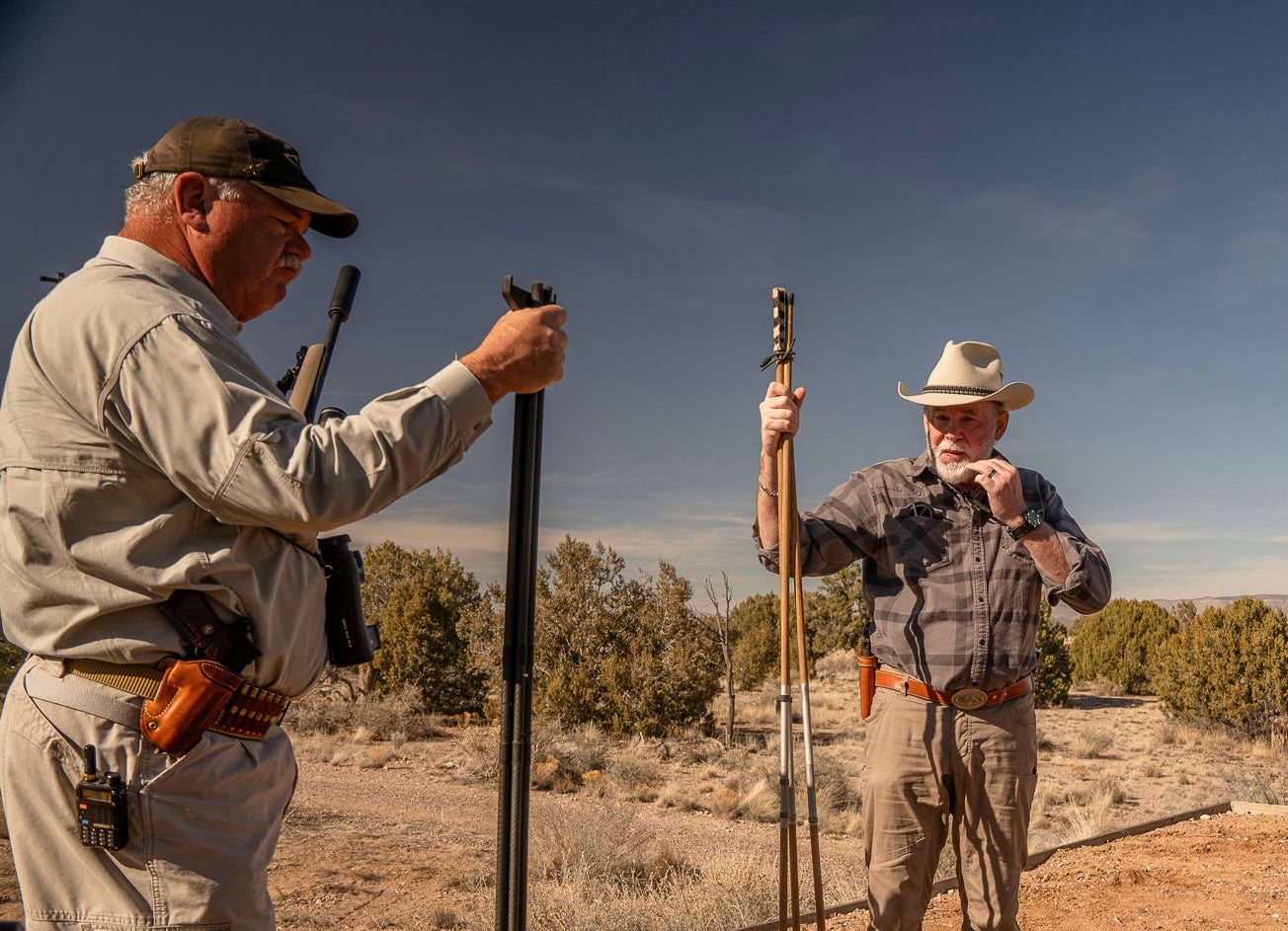 Shooting instructors stand at a gun range each holding tripod shooting sticks.