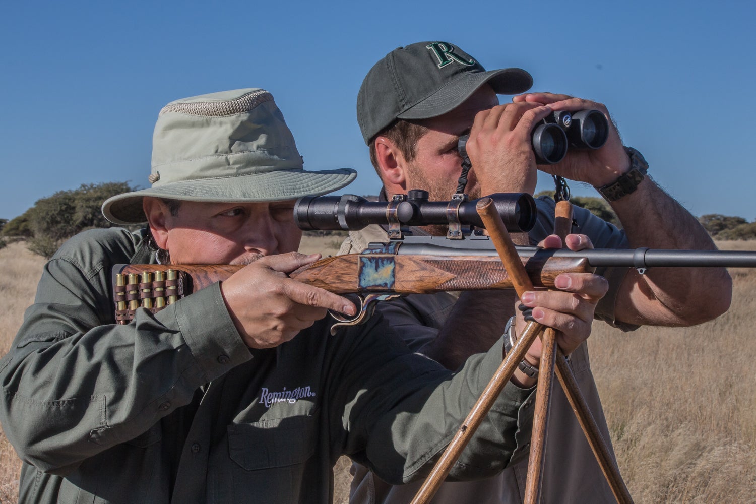 A hunter fires a rifle off shooting sticks in Africa, as his guide watches through binoculars