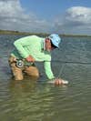 A fly fisherman in a green shirt and blue hat releases a bonefish in the water