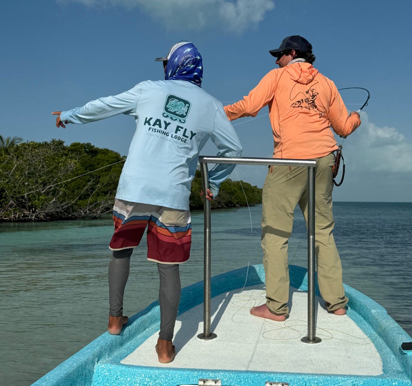 A fly fisherman in an orange shirt stands next to a fishing guide in a blue shirt on a skiff.